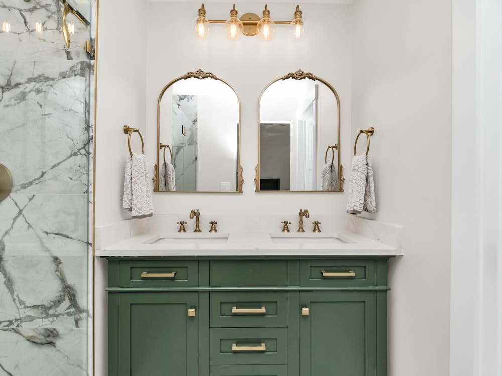 A bathroom with a green vanity, marble counter and arched mirrors